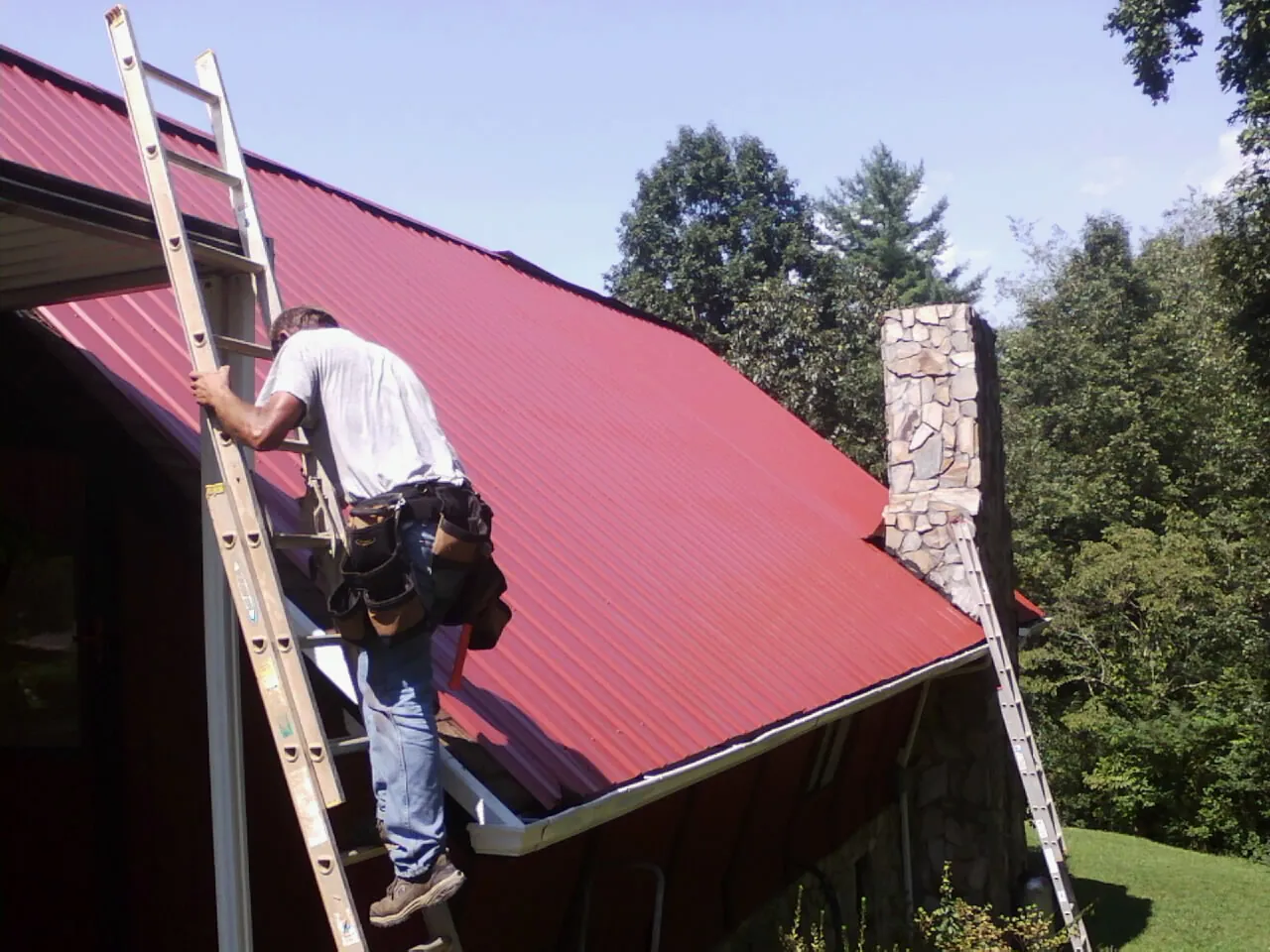 Roofer on a ladder beside a red metal roof and stone chimney — Soffit & Fascia Repair by Roofing Brothers in Toughkenamon